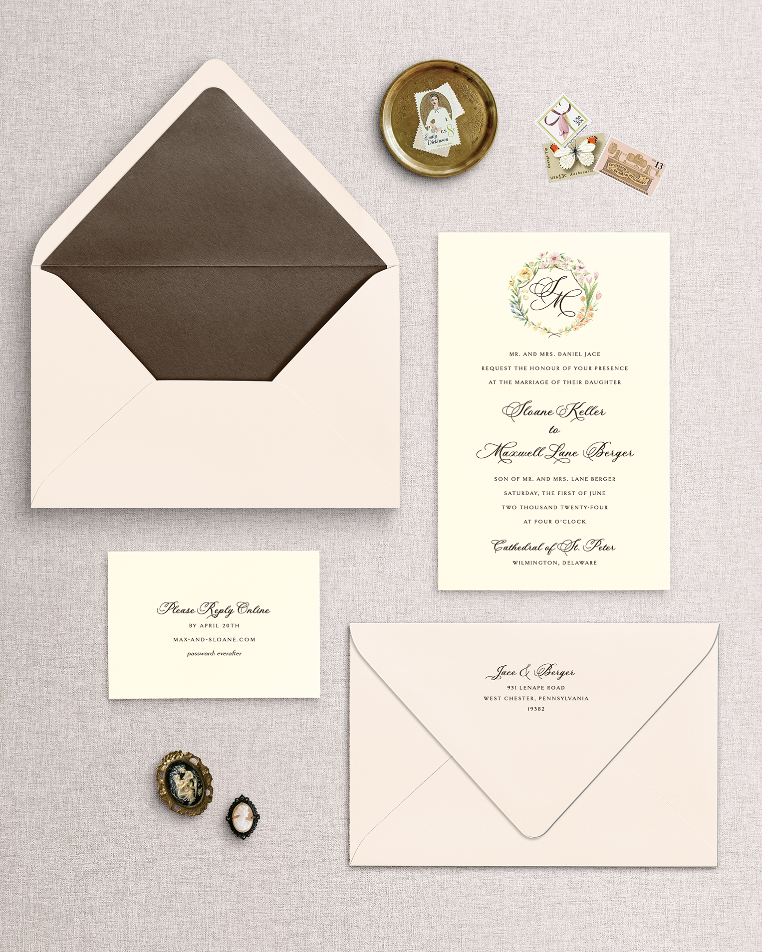 Wedding invitation set with envelopes and decorative items on a textured surface. Design includes ivory cards and a blush envelope. The invitation has a floral monogram crest at the top.